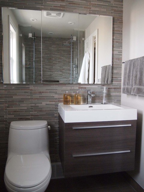 Mur sur lequel est installé un lavabo avec meuble en bois, des toilettes et un grand miroir. Des carreaux dans différentes nuances de brun et de gris.